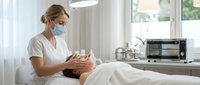 Professional female aesthetician in medical mask performing facial treatment on client in modern spa clinic with natural lighting and sterile equipment.