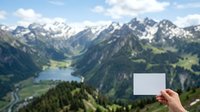 Unrecognizable person holding blank white card against alpine valley landscape with snow-capped peaks, forested slopes, and mountain lake for mockup purposes.
