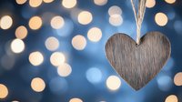 Wooden heart ornament hanging against blurred bokeh lights background in blue and gold tones, creating romantic holiday atmosphere.