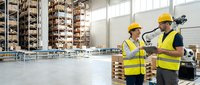 Two warehouse workers in safety vests discussing logistics in modern distribution center with shelving systems and natural lighting.