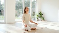 Young Asian woman in white clothing practicing meditation in lotus position on yoga mat in bright modern room with large windows and natural light.