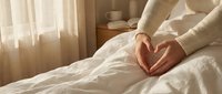 Woman making heart shape with hands on white bedding in cozy bedroom with warm morning sunlight streaming through curtains creating peaceful atmosphere.