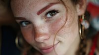 Young redhead woman with freckles and natural makeup looking at camera with soft smile and messy hair strands across face for beauty and lifestyle content.