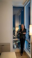 Confident businesswoman in black suit standing by floor-to-ceiling windows with city lights view in modern office interior for corporate success concepts.