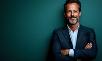 Confident mature businessman with gray beard wearing navy suit and light blue shirt, arms crossed, smiling against dark green background.