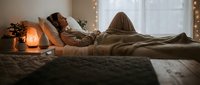 Woman with long brown hair lying on bed in cozy bedroom with warm lighting, salt lamp, and holiday decorations for relaxation and wellness content.