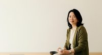 Asian woman in olive cardigan holding coffee cup, sitting at wooden table against beige wall, professional portrait for business and lifestyle content.