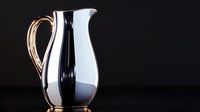 Elegant white ceramic pitcher with golden handle on black background, dramatic lighting creates reflective surface and sophisticated mood for luxury tableware styling.