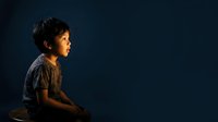 Young boy with dark hair and olive complexion, wearing brown shirt, sitting in profile against dark blue background with warm side lighting for portrait and child development concepts.