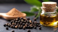 Black peppercorns, ground pepper powder and essential oil in glass bottle with cork on dark background with green leaf, macro food photography.