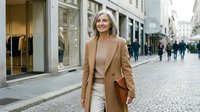 Confident mature blonde businesswoman walking on cobblestone street wearing camel coat and turtleneck, carrying leather bag in urban shopping district.