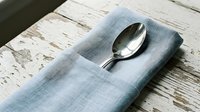 Silver spoon resting on folded blue linen napkin over rustic whitewashed wooden table surface for dining and tableware styling.