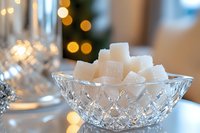 White sugar cubes stacked in crystal bowl with soft bokeh lights and festive holiday atmosphere in background, elegant table setting for special occasions.