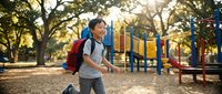 Young Asian boy with backpack running joyfully at colorful playground during golden hour sunlight filtering through trees creating warm outdoor recreation scene.