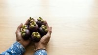 Hands holding fresh mangosteen fruits on wooden surface, tropical produce display for food and agriculture marketing materials.