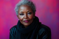 Elegant mature African American woman with silver hair and confident smile wearing black scarf against vibrant pink background, studio portrait capturing natural aging beauty.