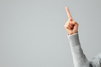 Raised index finger pointing upward against neutral gray background, showing attention or important point gesture in gray sweater sleeve.