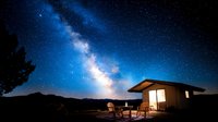 Glowing cabin under starry night sky with bright Milky Way galaxy, mountain silhouettes and empty chairs on porch creating cozy outdoor atmosphere.