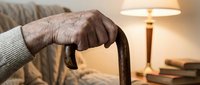 Elderly person's weathered hands gripping wooden walking cane in warm home interior with lamp and books, representing aging, mobility support, and independence.