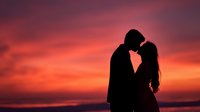 Romantic couple silhouette embracing against vibrant sunset sky with dramatic orange and pink clouds creating intimate moment for wedding and love concepts.