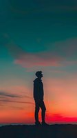 Silhouette of young man standing alone against dramatic sunset sky with vibrant orange and teal colors, capturing solitude and contemplation at dusk.