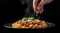 Hand sprinkling grated cheese over steaming pasta with meat sauce and fresh basil on dark plate against black background for culinary presentations.