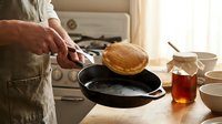 Caucasian male chef in apron flipping golden pancake in cast iron skillet on kitchen counter with stove and ingredients visible, cooking breakfast.