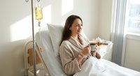 Young Caucasian woman patient in hospital bed with IV drip enjoying hot beverage during medical treatment recovery in bright healthcare facility room.