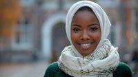 Young African woman wearing white hijab and green sweater smiling warmly at camera in urban setting with soft bokeh background for lifestyle content.