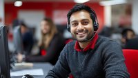 Young male customer service representative wearing headset smiling at camera in modern call center office with red interior and coworkers in background.