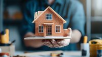 Small brick house model held in hands against blurred office background, representing real estate, property investment, and home ownership concepts.
