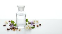 Clear glass bottle with essential oil surrounded by fresh herbs, flowers and seeds on white background for natural wellness and aromatherapy products.