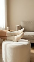 Cozy home relaxation scene with feet in white ribbed socks resting on ottoman, warm beige living room interior with coffee cup and natural lighting.