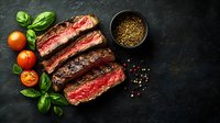 Juicy medium rare sliced beef steak with fresh basil leaves, cherry tomatoes, and peppercorn seasoning on dark stone background, top view food photography.