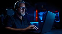 Middle-aged Caucasian man with gray beard wearing headset and glasses working on laptop in dark room with blue ambient lighting, focused on cybersecurity monitoring.
