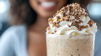 Creamy coffee milkshake topped with whipped cream and chocolate crumble, with smiling African American woman blurred in background.
