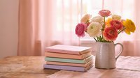 Pastel ranunculus flowers in a ceramic jug beside stacked colorful notebooks on a wooden table with soft pink curtains in background.