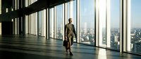 Professional businessman walking through modern office corridor with floor-to-ceiling windows overlooking city skyline during golden hour sunlight.