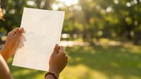 Person of African descent holding blank white paper outdoors in sunlit park setting, suitable for mockup, advertising, and promotional campaigns.