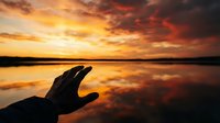 Hand silhouette reaching toward golden sunset over calm lake with dramatic orange and red clouds reflected in water creating peaceful spiritual moment.
