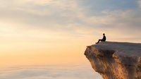 Silhouette of person sitting alone on cliff edge against dramatic sunset sky, contemplating vast horizon over peaceful ocean landscape.