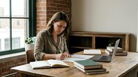 Young professional woman working at wooden desk in modern loft office with brick walls, writing notes while using laptop for business planning and productivity.