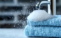 Fresh blue cotton towels stacked near running water tap with splashing droplets and foam, creating relaxing spa bathroom atmosphere for wellness and hygiene concept.
