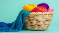 Colorful yarn balls in woven basket against turquoise background. Vibrant wool skeins in yellow, purple, red, pink and blue colors arranged for knitting and crafts.