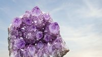 Large amethyst crystal cluster against a soft cloudy sky, showcasing natural purple gemstone formations for wellness and mineral themes.