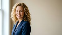 Professional blonde businesswoman with curly hair smiling confidently in navy blue suit jacket standing by office window for corporate headshot.