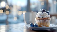 Fresh vanilla cupcake topped with whipped cream frosting and ripe blueberries on white plate against blurred bokeh background with warm cafe lighting.
