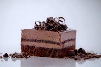 Decadent chocolate layer cake slice with mousse filling, topped with dark chocolate curls and chunks on a light background.