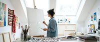 Young African American female artist painting on canvas in bright modern art studio with skylights and creative supplies scattered on desk.