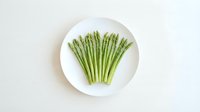Fresh green asparagus spears arranged on white ceramic plate against clean background for healthy cooking and nutrition concepts.